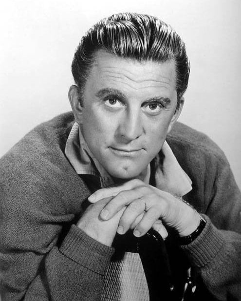Black and white portrait of a man with slicked-back hair, wearing a collared shirt and sweater, resting his chin on his folded hands and looking directly at the camera with a slight smile.