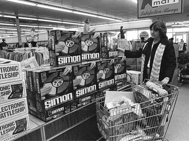 A person stands in a store aisle next to a display of stacked Simon electronic game boxes, with a shopping cart filled with items and shelves of clothes in the background.