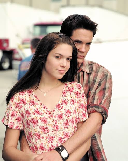 A young woman in a floral blouse stands in front of a young man wearing a plaid shirt. The man has his arms wrapped around her waist as they both look at the camera. The background is slightly blurred with outdoor elements.