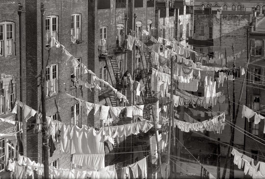 Rows of white laundry hang on clotheslines strung between brick apartment buildings with fire escapes, creating a dense web of linens in a city alley. The image is black and white, evoking a historical urban setting.