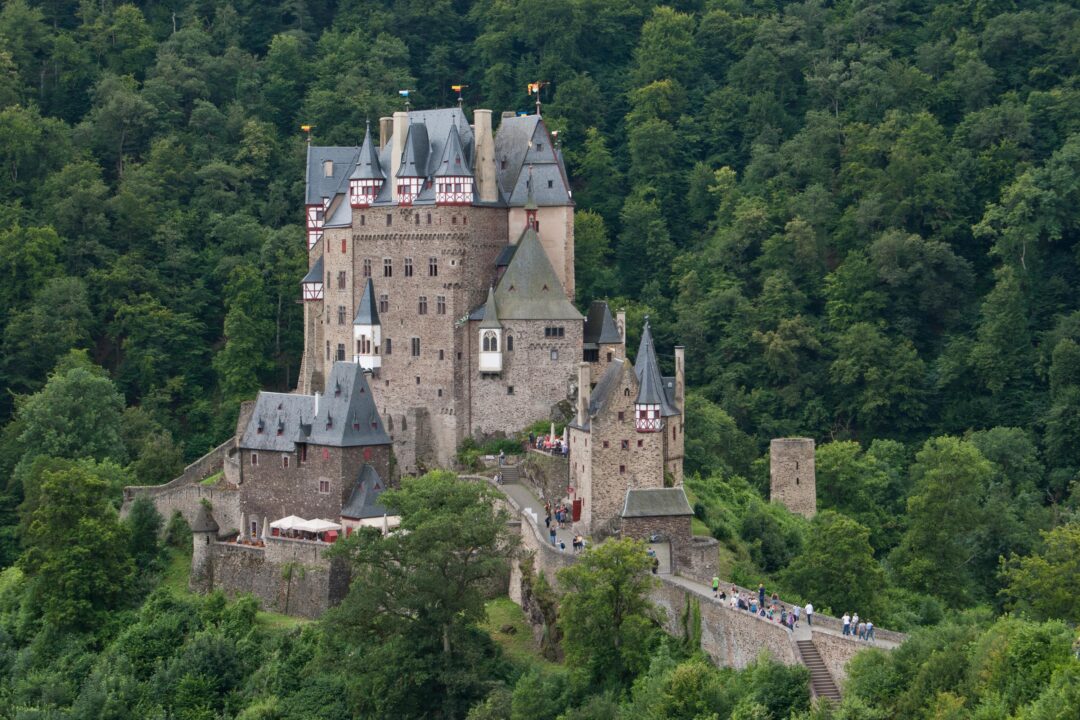A large medieval stone castle with pointed towers sits on a small hill, surrounded by dense green forest. People are walking along a stone bridge leading to the castle entrance.