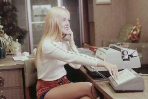 A woman with long blonde hair sits at an office desk, talking on a corded phone while using a typewriter. She wears a white long-sleeve top and red shorts. The office has vintage decor and green furniture in the background.