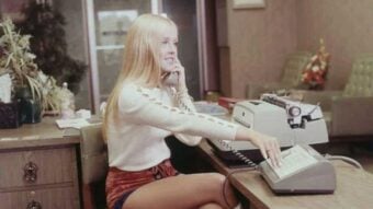 A woman with long blonde hair sits at an office desk, talking on a corded phone while using a typewriter. She wears a white long-sleeve top and red shorts. The office has vintage decor and green furniture in the background.