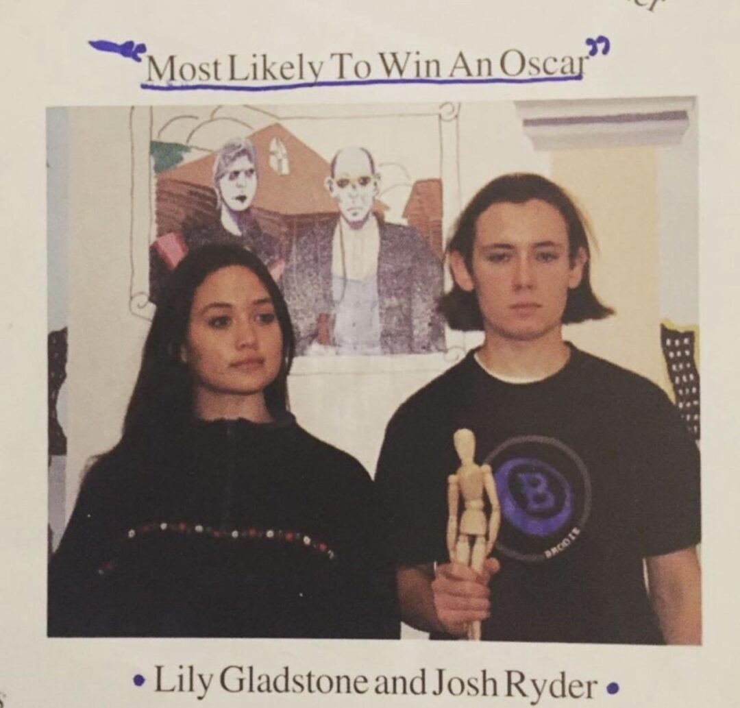 Two young people stand side by side in front of an art piece. The person on the right holds a wooden mannequin. Text above reads “Most Likely To Win An Oscar,” and below are the names Lily Gladstone and Josh Ryder.