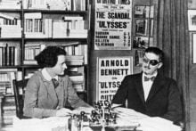 A woman and a man in dark glasses and a bow tie sit at a desk in a book-filled room, with posters promoting &ldquo;Ulysses&rdquo; and other books visible in the background.