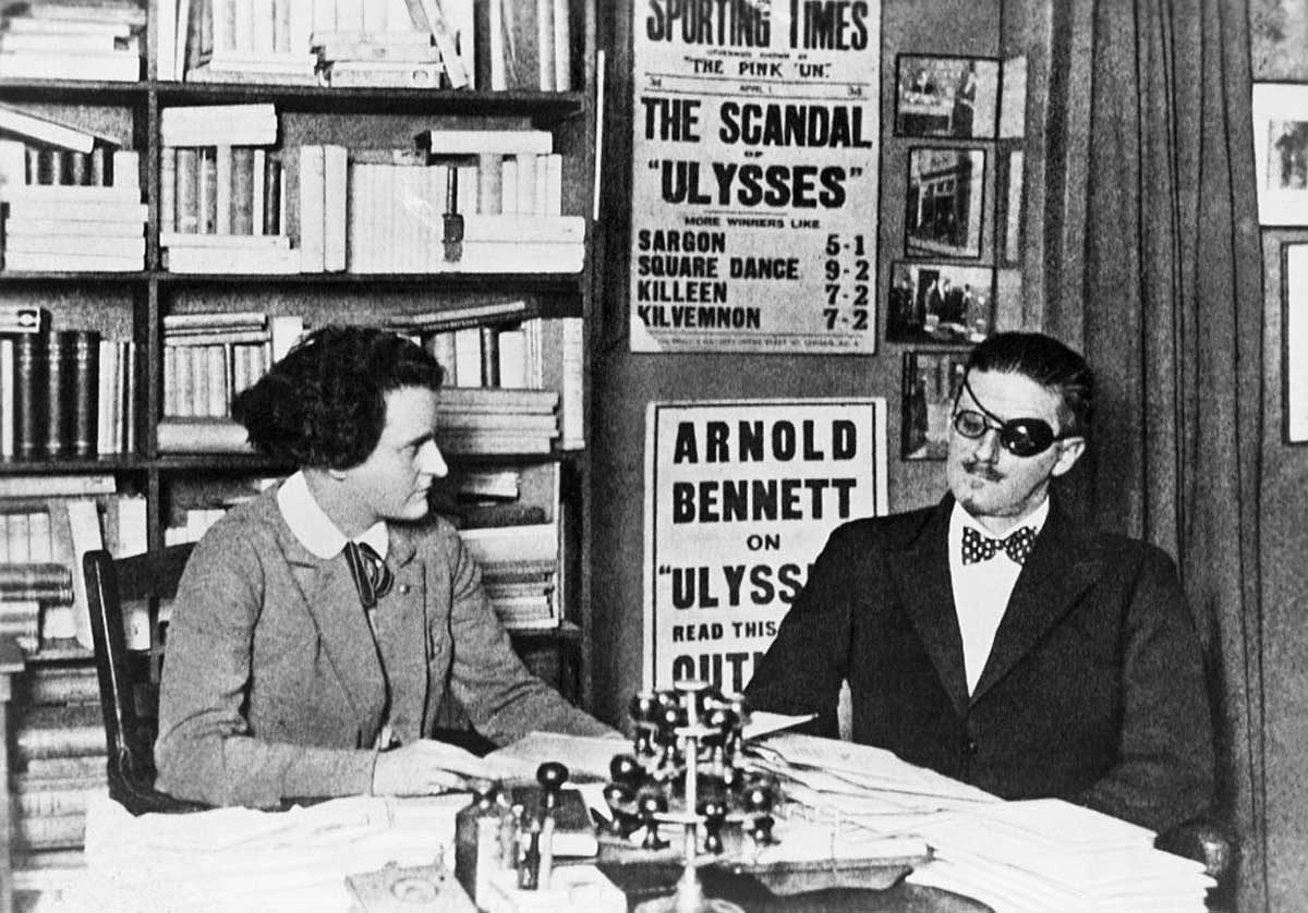 A woman and a man in dark glasses and a bow tie sit at a desk in a book-filled room, with posters promoting “Ulysses” and other books visible in the background.