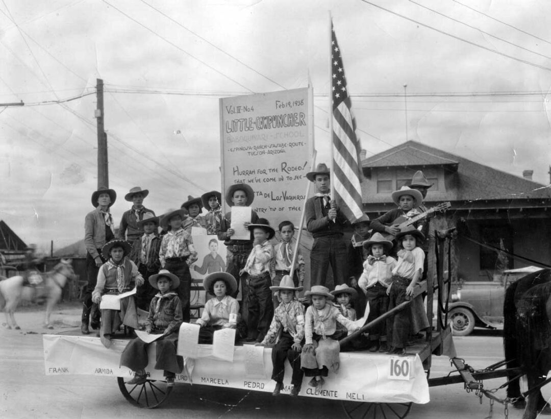 A black-and-white photo of children and adults in cowboy hats and western attire on a parade float with an American flag, a large sign, and a backdrop of buildings and power lines.