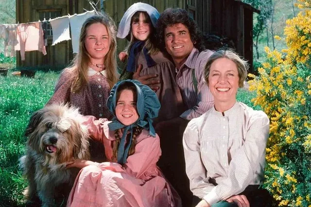 A smiling family in old-fashioned prairie clothes poses outdoors with a dog, near a wooden house and laundry line. Yellow flowers and greenery surround them, evoking a rural, historical setting.