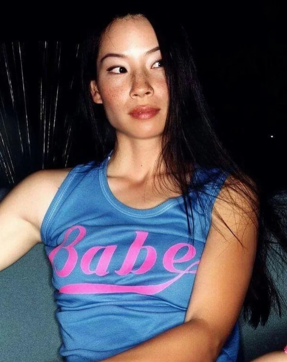 A woman with long dark hair sits on a couch, wearing a blue sleeveless top with the word "Babe" written in pink script. She is looking to her left with a neutral expression.