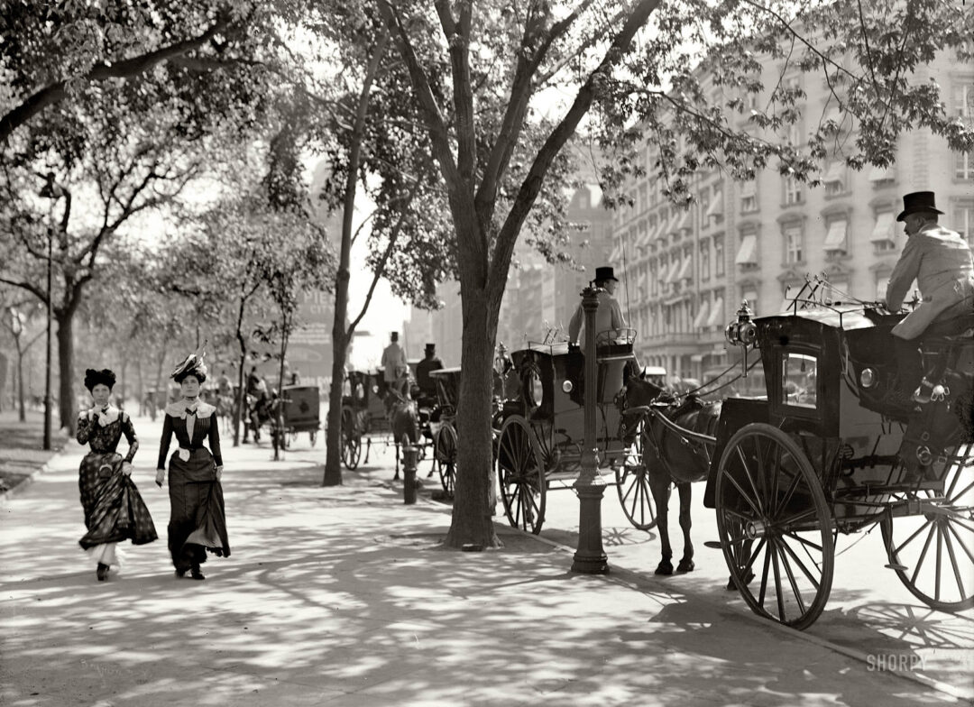 Two women in Victorian-era dresses walk along a tree-lined city street, passing several horse-drawn carriages. The background shows ornate buildings and more people and carriages, evoking a bustling late 19th-century urban scene.