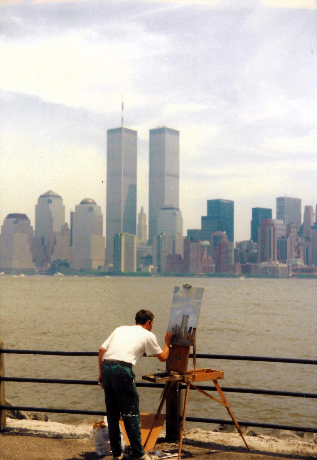 A person paints the New York City skyline, including the Twin Towers, on an easel by the waterfront. The river and a cityscape with tall buildings are visible in the background.