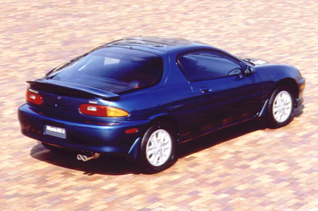 A dark blue, two-door sports coupe parked on a brick surface, viewed from the rear left side. The car has a smooth, aerodynamic design, tinted windows, and a subtle rear spoiler.
