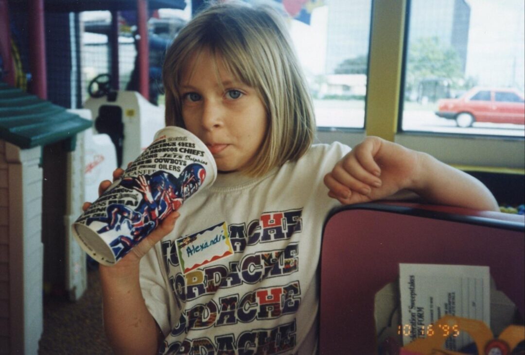 A young girl with blonde hair drinks from a large cup with a sports design. She wears a white graphic T-shirt and a name tag that says "Alexandra." She is indoors, sitting beside a counter, with a window and cars outside.