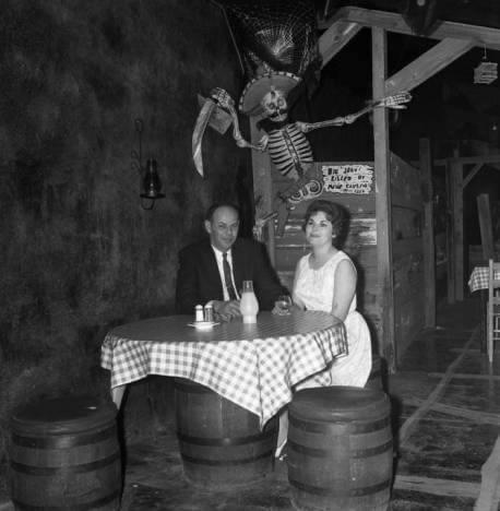 A man in a suit and a woman in a dress sit at a table covered with a checkered cloth, using barrels as seats. Behind them, a skeleton pirate decoration with a sword hangs on the wall.