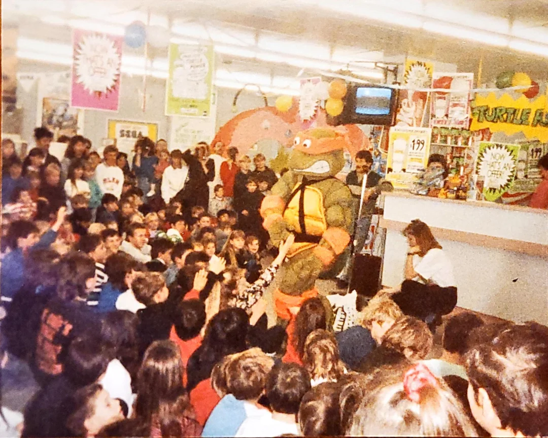 A person in a Teenage Mutant Ninja Turtles costume entertains a large crowd of children and adults inside a brightly decorated store filled with colorful posters and shelves of toys.