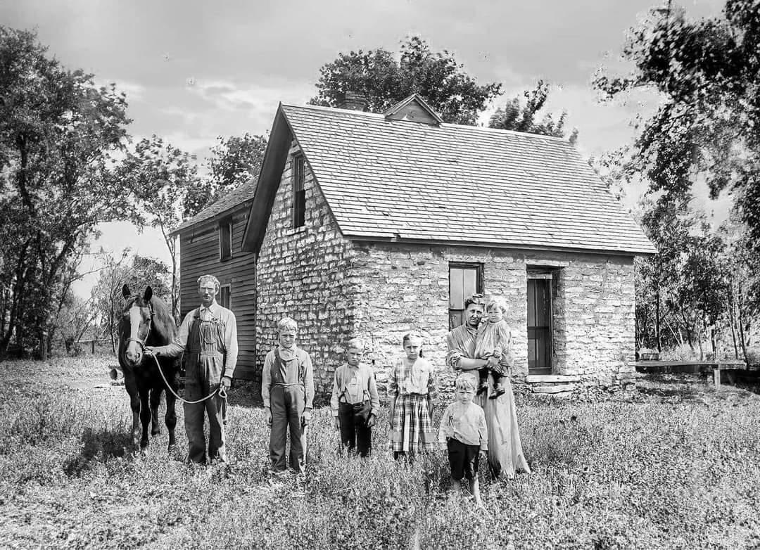 A black-and-white photo shows a family of seven standing in front of a small stone house. The group includes two adults, five children, and a horse, with trees and grass surrounding the scene.