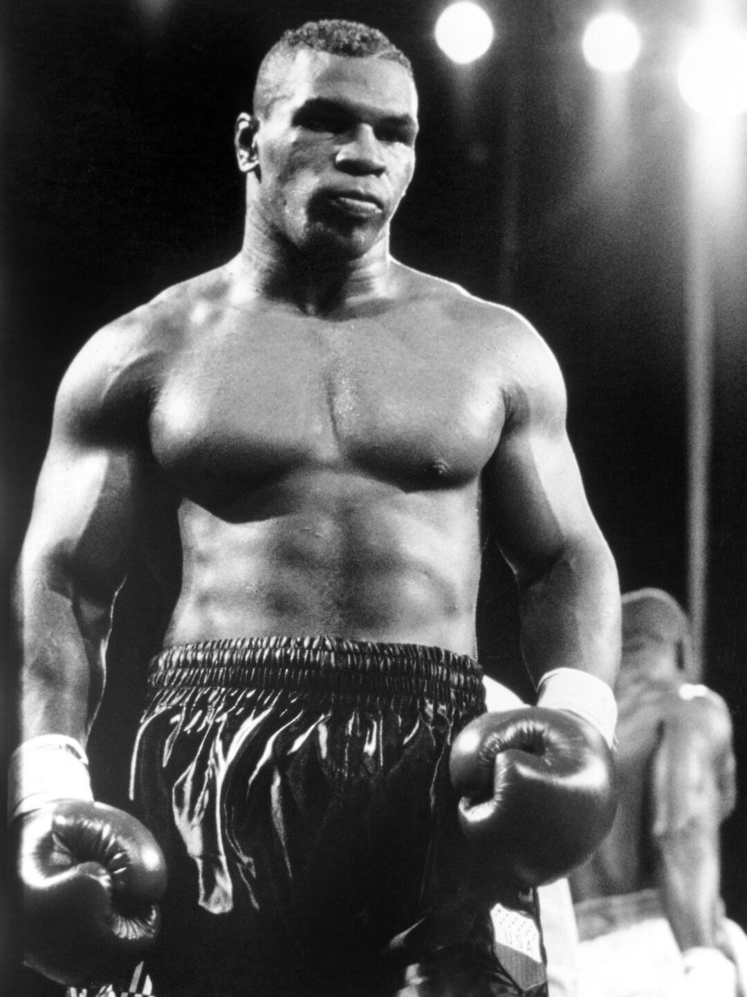 A muscular male boxer stands confidently in a boxing ring, wearing dark boxing shorts and gloves, with bright lights shining overhead and another boxer blurred in the background. The image is in black and white.
