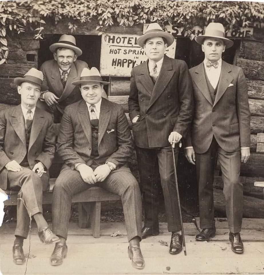 Five men in suits and hats pose in front of a rustic wooden building with a sign reading “Hotel Hot Springs in Happy.” Two are seated on a bench, three stand behind them, all looking at the camera.