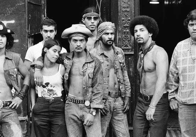A group of eight young adults pose together in front of a worn building, wearing casual and punk-inspired clothing, denim vests, and accessories. Some look directly at the camera, while others appear relaxed.