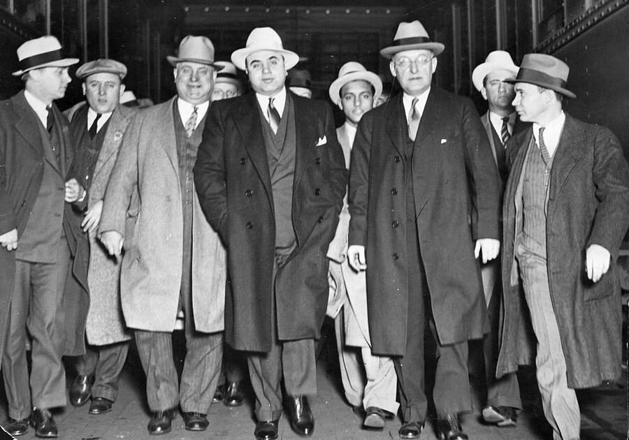 A group of men in suits and hats walk together in what appears to be a train station or industrial setting. The central figures look serious, dressed in dark overcoats, while others surround them, also wearing hats and coats.