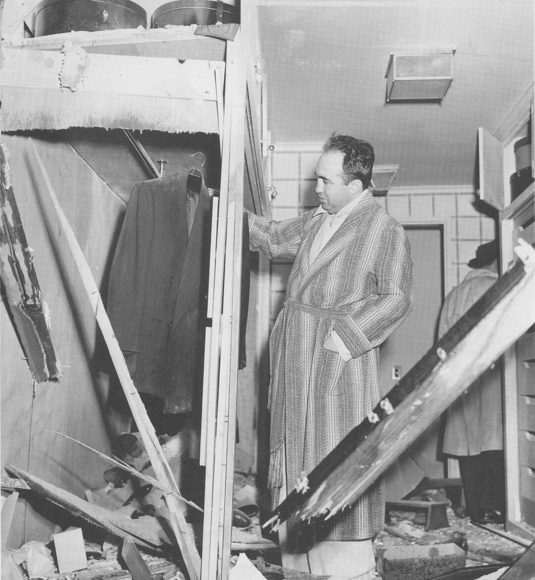 A man in a bathrobe looks at a closet in a heavily damaged room with debris, broken wood, and a torn ceiling. Another person stands in the background. The scene appears to be indoors, possibly after an explosion or disaster.