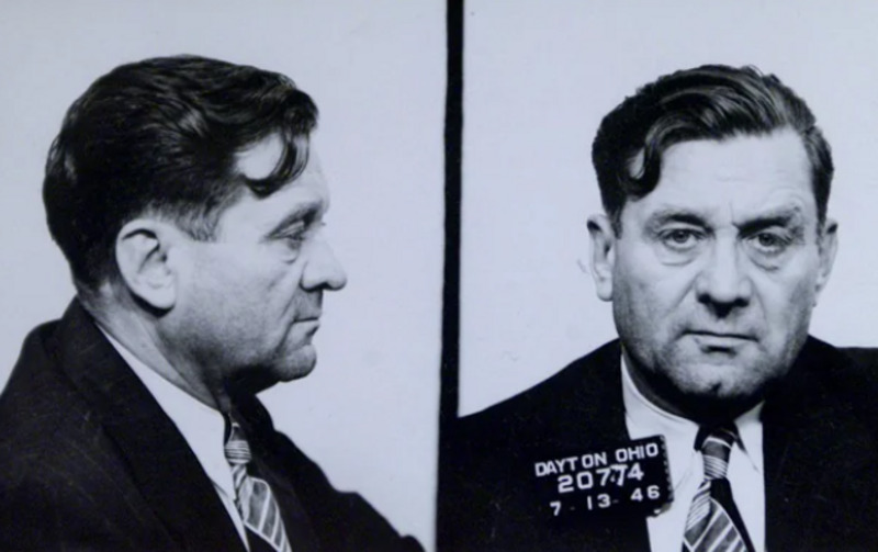 Black and white mugshot of a man in a suit and tie, shown in both profile and front views. He has wavy dark hair, and the sign reads "Dayton Ohio 20774, 7-13-46.
