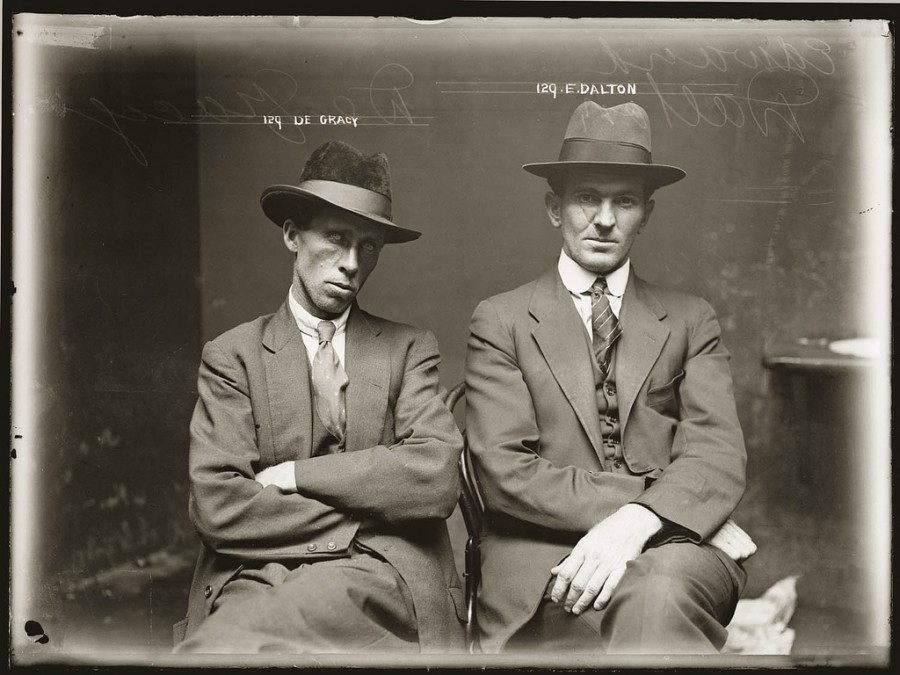 Two men in suits and hats sit side by side against a dark backdrop. One sits with arms crossed and a tilted head; the other sits upright with hands on his lap. Both stare seriously at the camera.