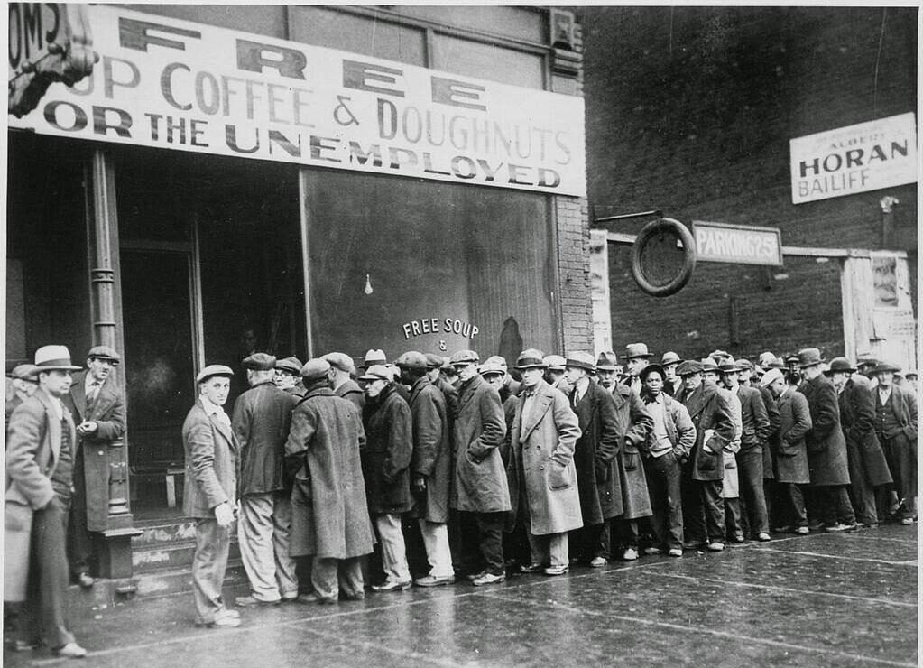A long line of men in coats and hats wait outside a building with a sign reading "Free Coffee & Doughnuts for the Unemployed." The scene appears to be from the Great Depression era.