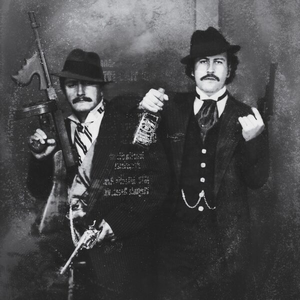 Two men in vintage suits and hats pose with stern expressions; one holds a Tommy gun, the other holds a bottle of Jack Daniel's whiskey and shows a middle finger. The black-and-white photo has an old, grainy effect.