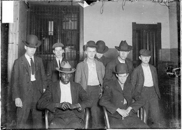A black-and-white photo of eight young men in suits and hats. Two are seated in front, while six stand behind them. They are indoors near barred doors or windows, posing seriously for the camera.