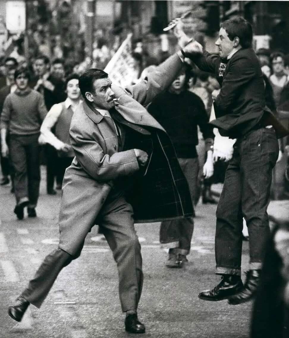 A man in a long coat punches another man in midair on a busy street, while a crowd of people watch the confrontation in the background.