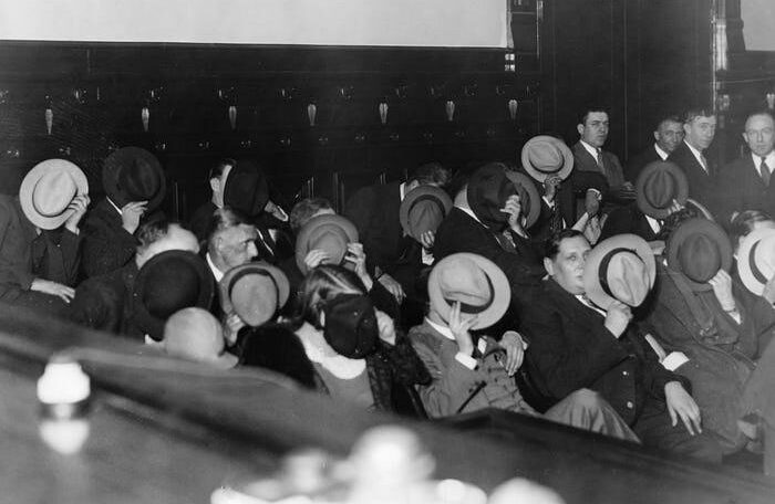 A group of men in suits sit closely together in a courtroom, hiding their faces behind hats or hands, while a few others stand in the background observing.