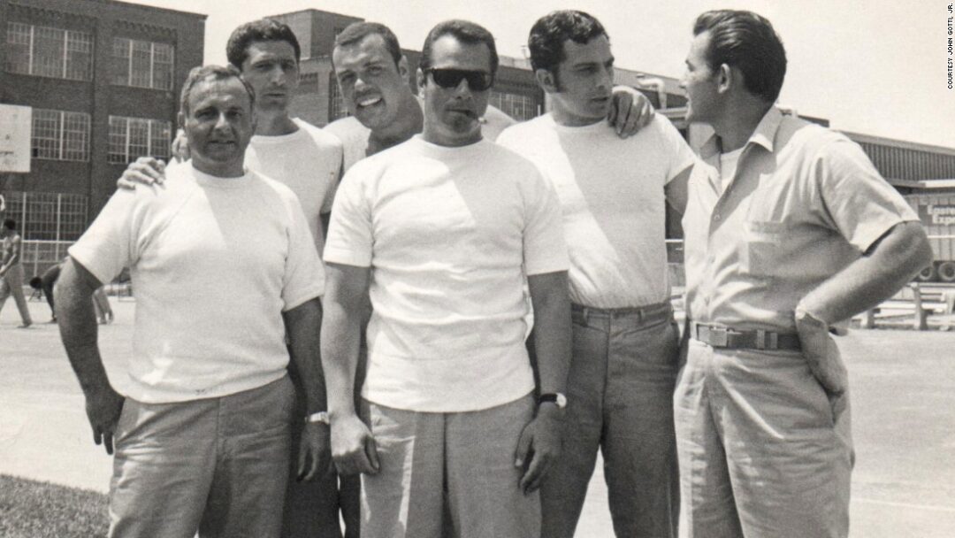 Six men stand together outdoors in casual clothing, posing and smiling for the camera on a sunny day, with buildings visible in the background.