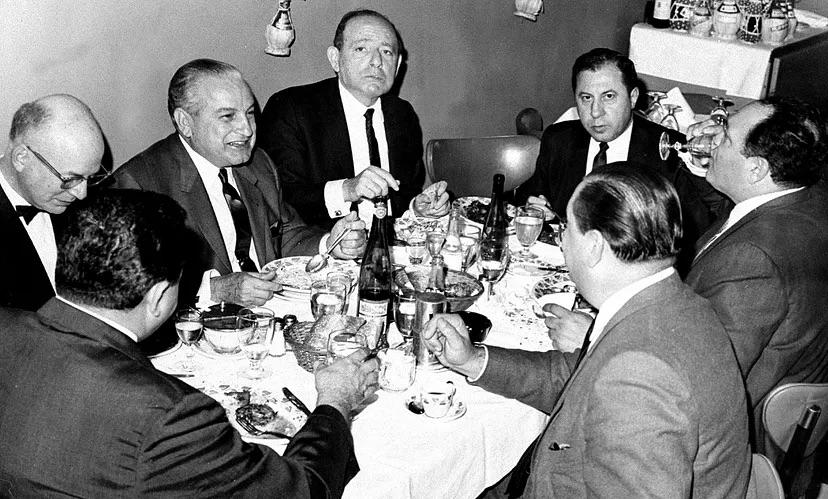 Seven men in suits sit around a table set with food, drinks, and wine bottles, engaged in conversation during a formal meal in a black-and-white photograph.