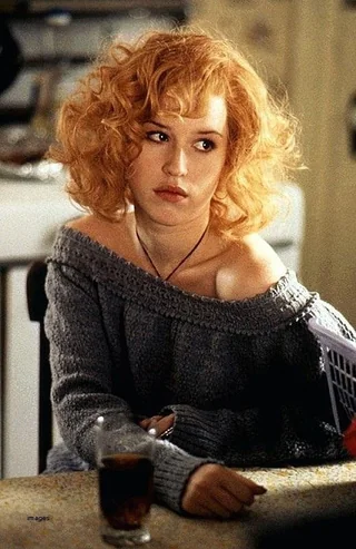 A young woman with curly red hair sits at a table wearing an off-the-shoulder gray sweater. She looks to the side with a thoughtful expression, resting her arm on the table beside a glass of dark liquid.