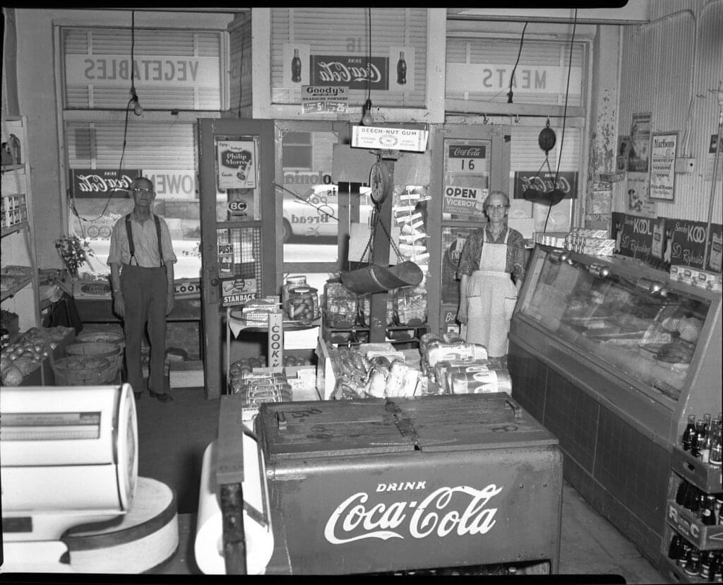 Black and white photo of an old-fashioned grocery store. Two people stand behind counters filled with goods. A Coca-Cola cooler is in front, with signs for meats and vegetables visible in the background.