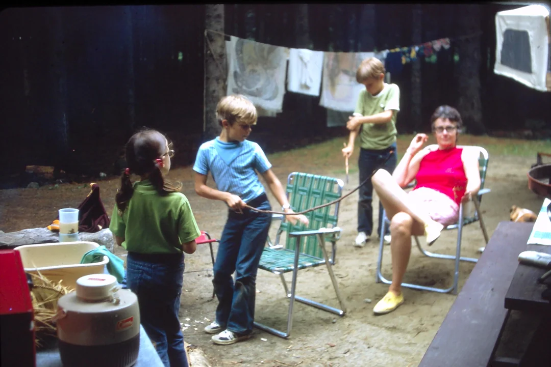 Three children play outdoors with a rope near an adult sitting in a lawn chair. Laundry hangs on lines in the background, and outdoor items are scattered around the yard. The scene appears relaxed and casual.