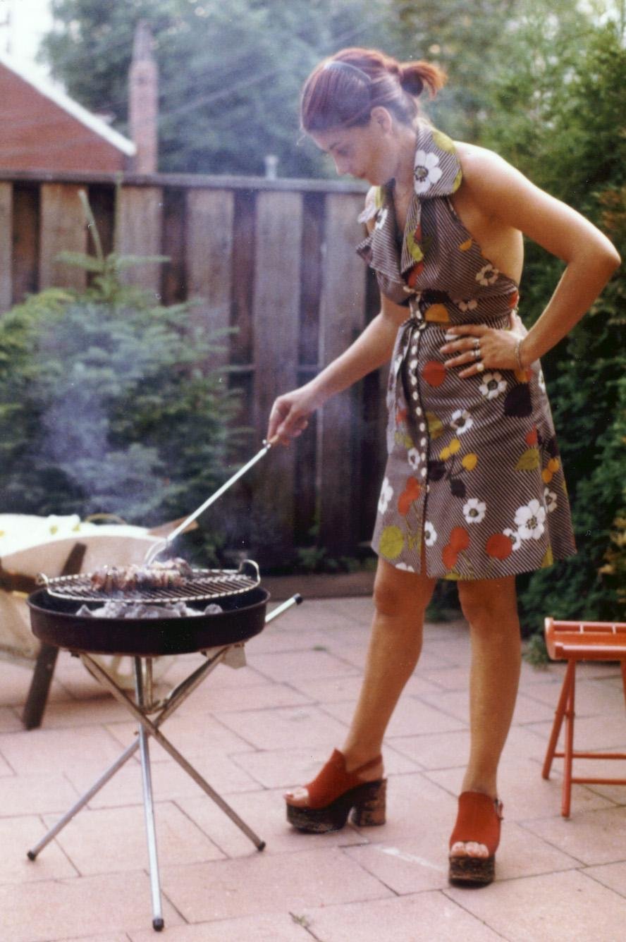 A woman in a floral dress and red platform sandals grills food on a small barbecue in a backyard patio, surrounded by greenery and a wooden fence. Smoke rises from the grill as she tends to the food.