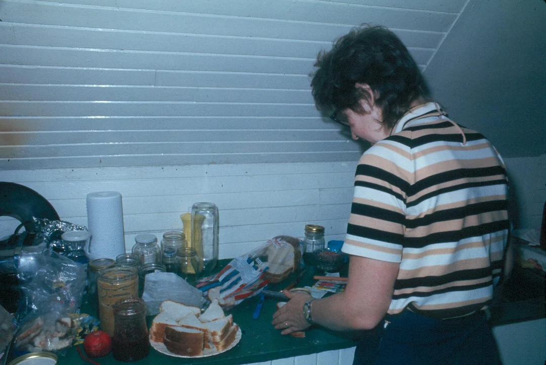 A person in a striped shirt stands at a kitchen counter, preparing food. Sliced bread, jars, and other ingredients are spread out on the green counter under a sloped white ceiling.