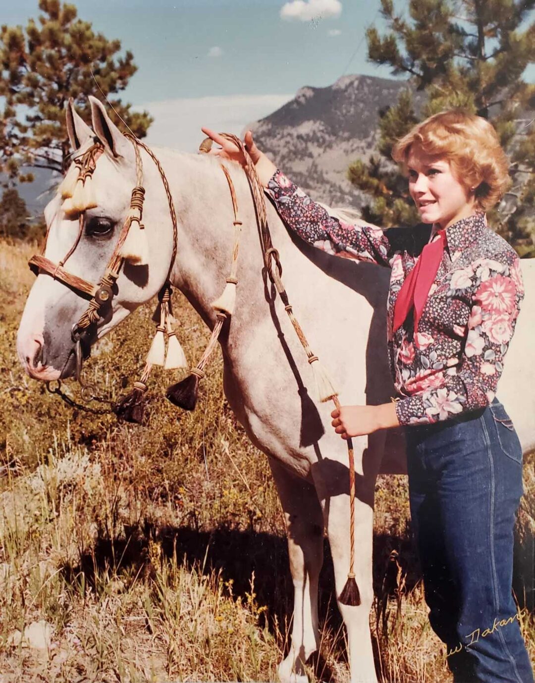 A young woman in a floral shirt and jeans stands outdoors on a grassy hillside, holding the reins of a white horse with decorative tack. Pine trees and a mountain are visible in the background under a clear sky.