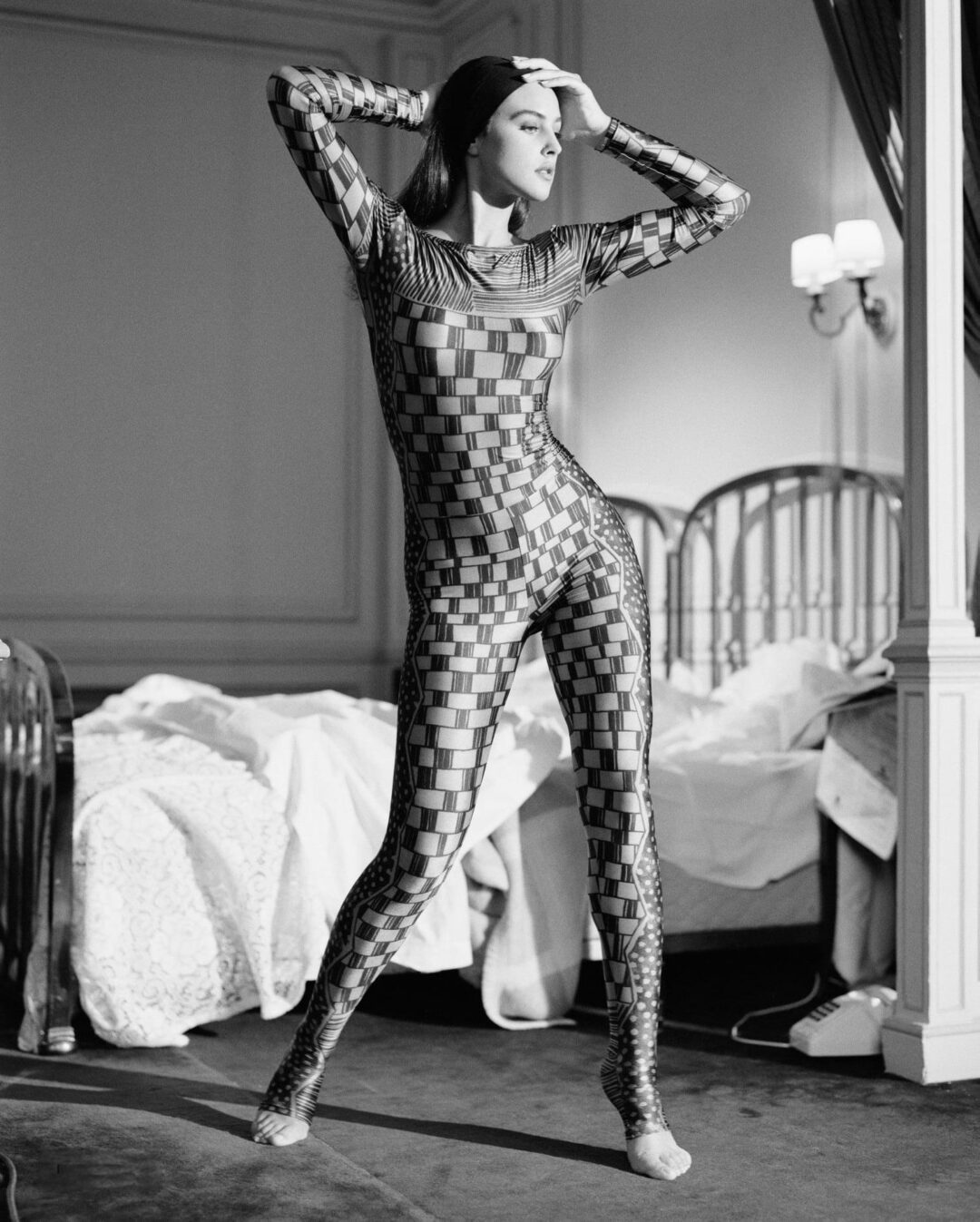 A woman stands in a bedroom wearing a fitted, geometric-patterned bodysuit. She poses with one hand on her head and the other on her hip. The room has unmade beds and classic wall decor, in black and white.