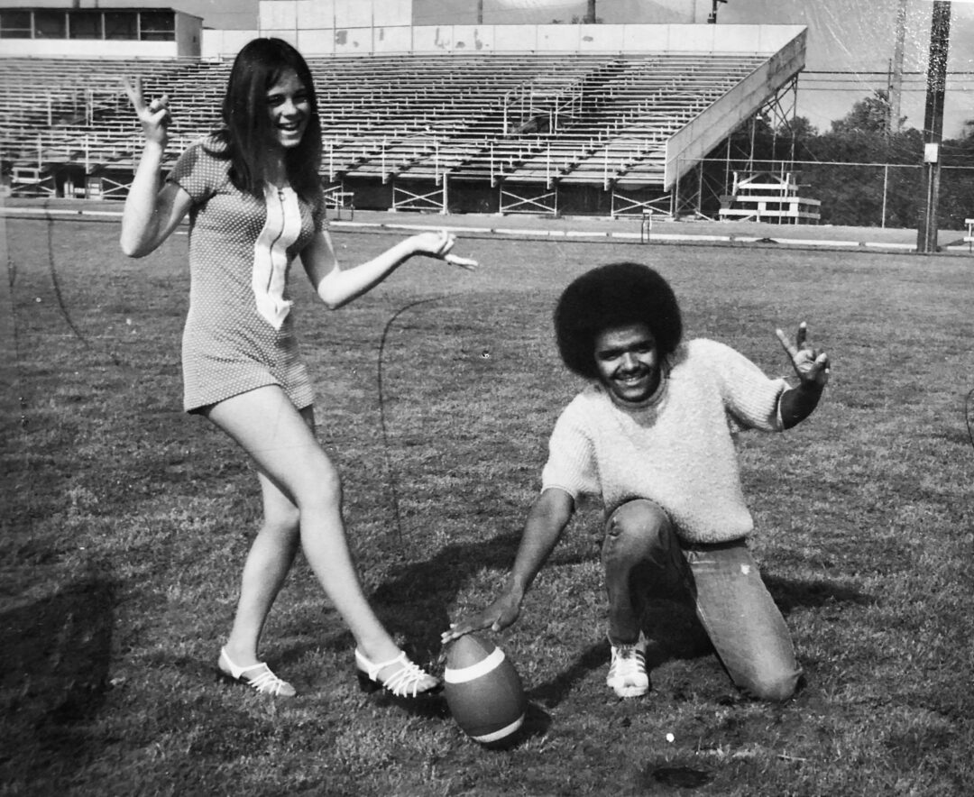 A young woman and a young man pose on a football field; the man kneels and holds a football upright while the woman stands beside him, both smiling and making peace signs. Empty bleachers are visible in the background.