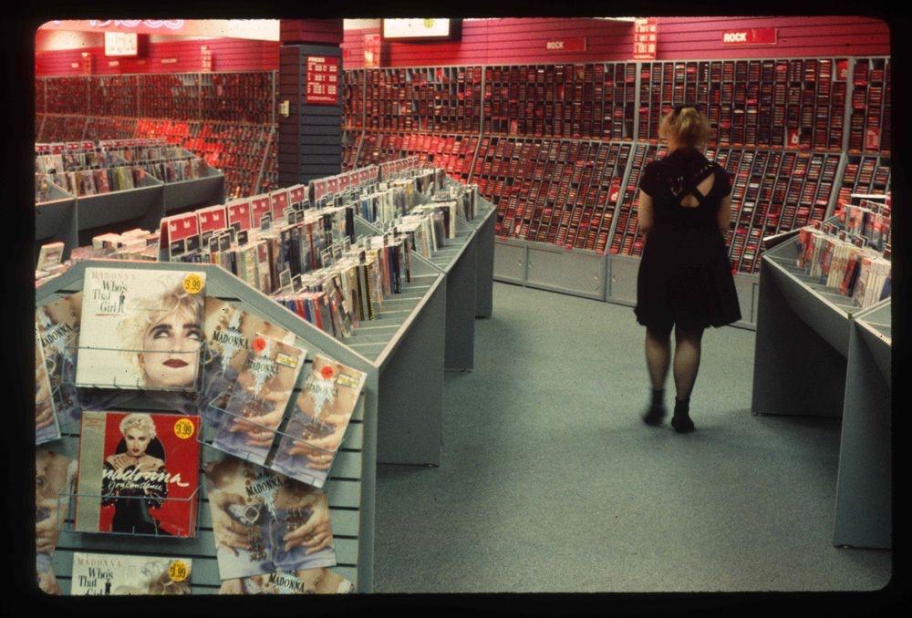 A woman in a black dress walks through a 1980s or 1990s music store filled with CDs and cassette tapes; albums by Madonna are prominently displayed in the foreground.