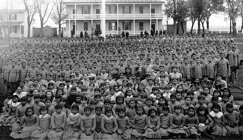 Black and white photo of hundreds of children, mostly girls in front rows and boys in back, posing in uniform rows outside a large two-story building with a porch and trees around it.