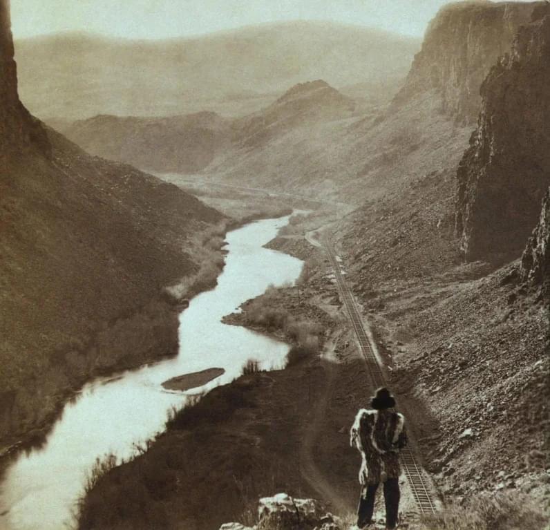 A person in a hat and coat stands on a rocky cliff overlooking a winding river and a railway track running through a deep canyon with high rock walls and distant mountains in the background.