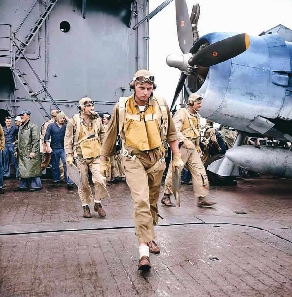A group of World War II-era pilots in tan uniforms and life vests walk with purpose on an aircraft carrier deck beside a blue propeller plane, while crew members and ship equipment are visible in the background.