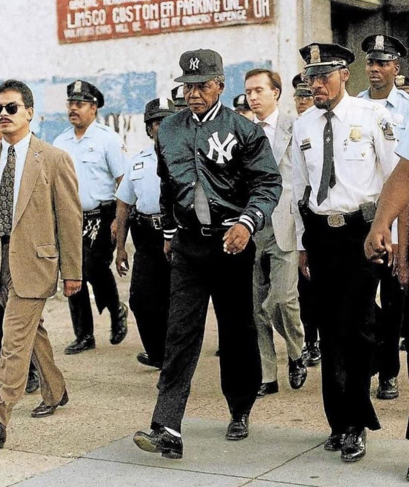 A man in a Yankees jacket and cap walks surrounded by several police officers and others in suits on a city street, with a faded building and sign in the background.