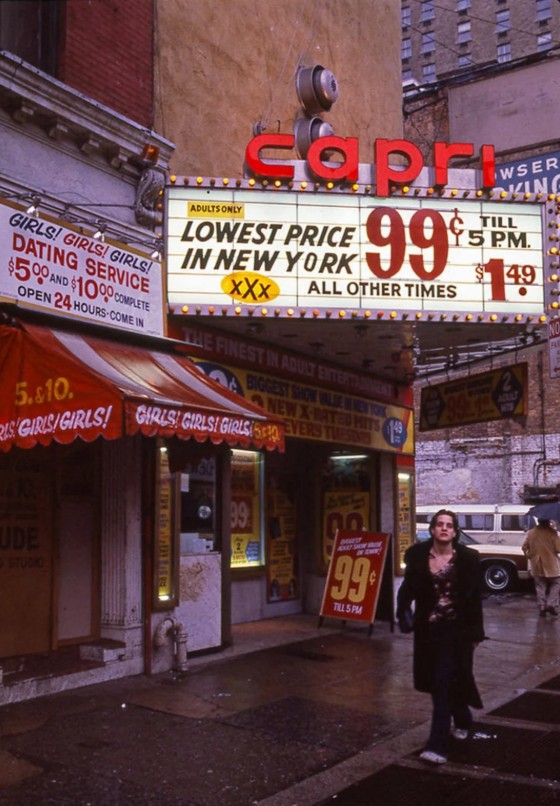 A person walks past the Capri theater in New York, with a bright marquee advertising adult movies for 99¢ until 5 p.m. Signs promote a dating service and "Girls! Girls! Girls!" The street is wet, and buildings are visible in the background.