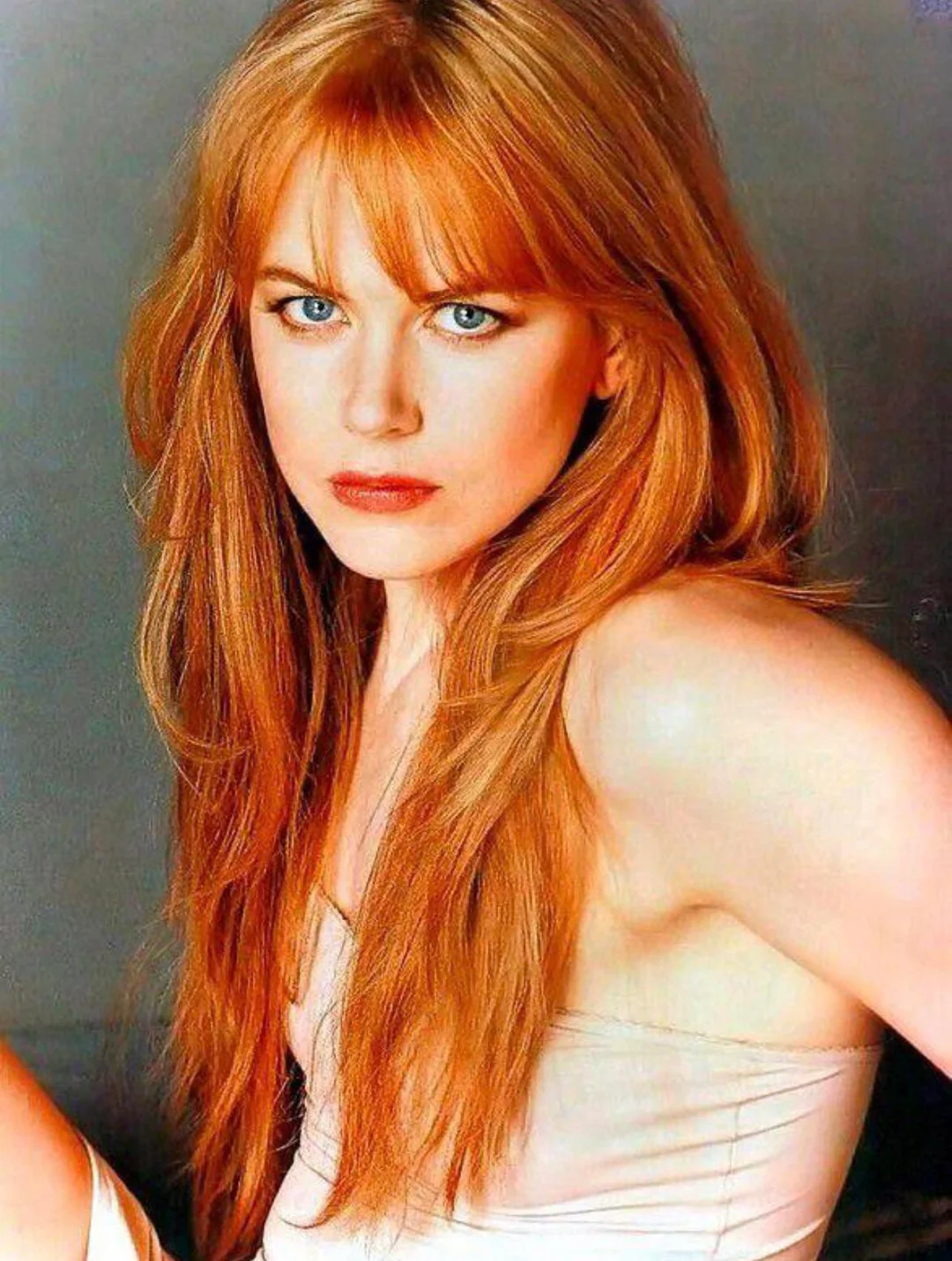 A woman with long red hair and bangs, wearing a light-colored sleeveless top, looks directly at the camera with a serious expression against a plain background.