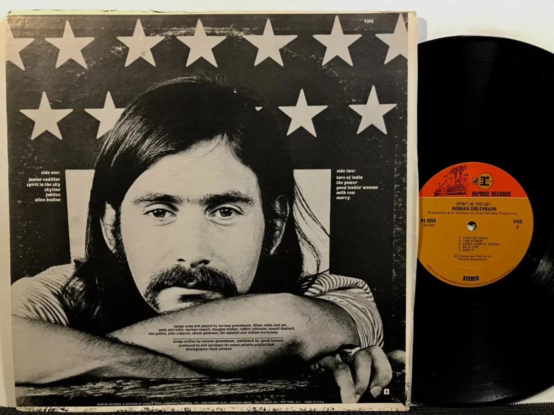 Black-and-white photo of a man with long hair and a mustache resting his arms on a table, face forward, against a backdrop of stars; a vinyl record with an orange label is beside the album cover.